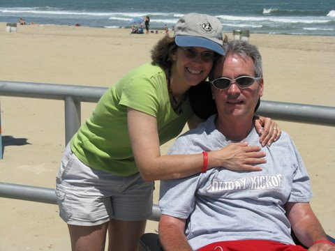 The smiling AJ has her arms wrpped around her always joful husband Jeff, already in a wheelchair, outside in front of the oceanfront with the sun at their backs. 