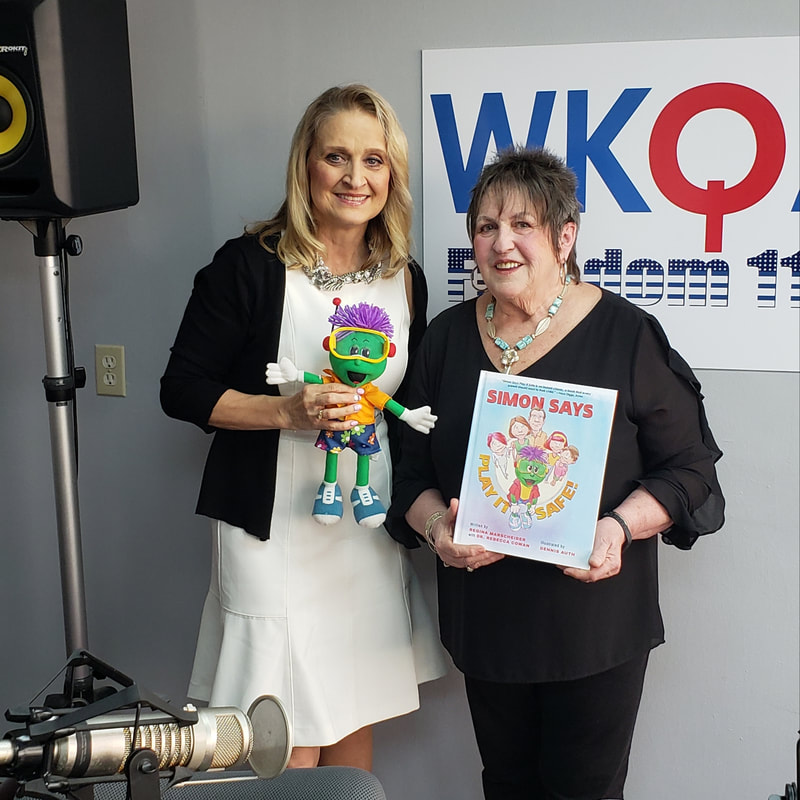 Dr. Christine Bacon in the WKQA studios holding the green and purple Simon doll while Regina, standing at her side, proudly displays her book 