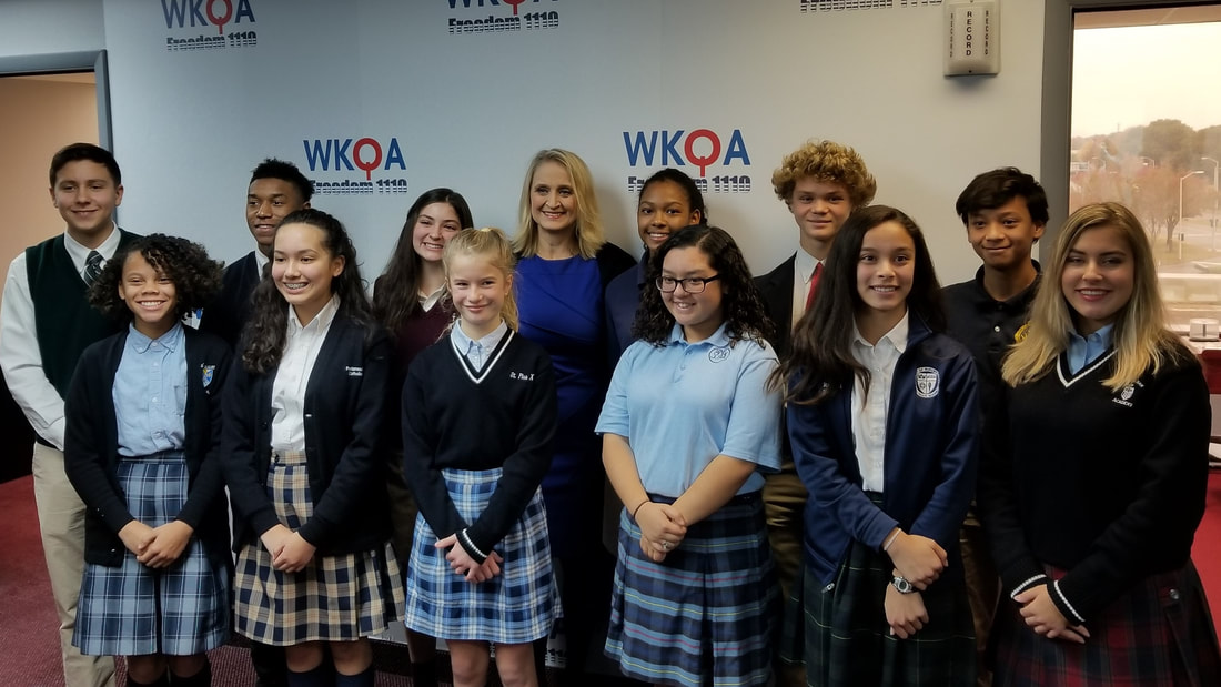 Dr. Christine Bacon standing amidst eleven uniform clad Catholic school students in the WKQA studios.